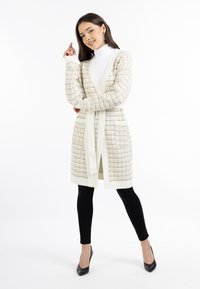 Texturierte, cremefarbene Strickjacke mit Gürtel und offener Front; hat ein Strickmuster und lange Ärmel. Kombiniert mit schwarzen Leggings und High Heels.