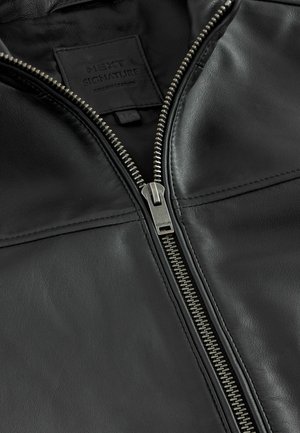 Black leather jacket featuring a silver zipper, textured surface, and a label sewn into the inside with the brand name "NEXT SIGNATURE."