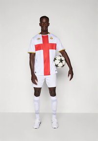 Bela nogometna majica z rdečim križem in zlatimi poudarki, kombinirana z belimi kratkimi hlačami in nogavicami. Igralec drži črno-belo nogometno žogo.