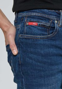 Jean en denim bleu foncé avec une texture lisse, doté d'une étiquette rouge sur la poche arrière et d'accents en métal argenté.