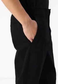 Fekete pamut nadrág, oldal zsebbel, egyenes lábformával és látható varrási részletekkel. Az anyag sima tapintású.