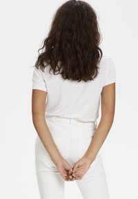Mujer con cabello rizado oscuro que lleva una camiseta blanca y pantalones blancos, de pie de espaldas, con las manos entrelazadas detrás de la espalda.
