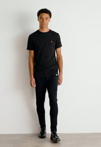 Tricou negru cu mânecă scurtă și un mic logo roșu, purtat cu pantaloni negri slim și pantofi negri, pe un fundal simplu deschis.