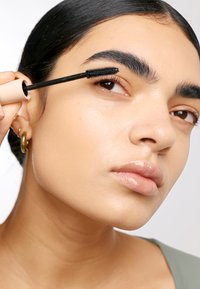 Mascara borste med svarta borst appliceras på en models mörka ögonfransar. Modellen har slät hy och en neutral grön topp.