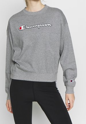 Persoon draagt een grijze Champion trui met logo op borst en mouw, gecombineerd met zwarte legging met witte zijstrepen.