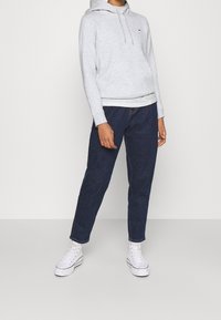 Grau Kapuzenpulli mit einer Fronttasche und Kordelzügen, kombiniert mit dunkelblauen Jeans und weißen Sneakern. Der Stoff wirkt weich und lässig.