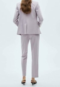 Costume violet clair sur mesure avec un blazer structuré et un pantalon à jambes droites. Tissu lisse avec un léger éclat. Escarpins noirs à bout pointu.