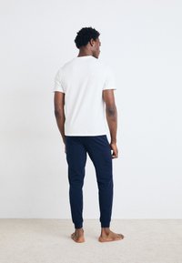 Tricou alb cu mânecă scurtă și pantaloni de jogger bleumarin, design ajustat, material moale, manșete ribbate, purtat desculț, văzut din spate pe un fundal simplu.