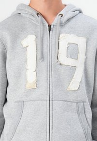 Siva zip-up zipka s ispucanim brojem "19" na prednjoj strani, s kengur džepovima i kapuljačom s čipkom.