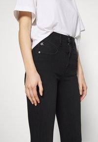 Czarne dżinsy z denimu o dopasowanym kroju, jasnym szwem, przednimi kieszeniami i małym logo CK na prawej kieszeni. Połączone z białym t-shirtem.