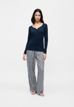 Femme portant un haut henley bleu marine à manches longues et un pantalon de pyjama à carreaux noir et blanc, debout pieds nus sur un fond clair uni.
