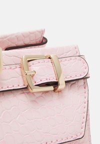 Bandoulière de sac à main texturée rose clair avec boucle rectangulaire dorée et couture ton sur ton sur une surface lisse.