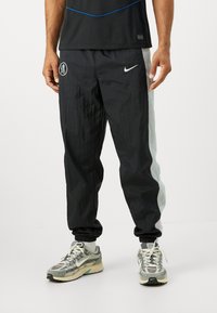Black track pants with a light gray side panel, made from a textured material. Features Nike logo and elastic cuffs.