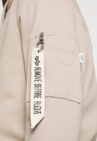 Beidzīgas krēmbalts džemperis audums ar sānu kabatu ar rāvējslēdzēju un baltu auduma birku ar uzrakstu "NOŅEMT PIRMS LIDOJUMA", kas piestiprināta ar klipu.