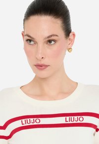 Maglione bianco con righe orizzontali rosse e testo "LIUJO", con scollo rotondo. Orecchini a forma di cuore dorati completano il look.
