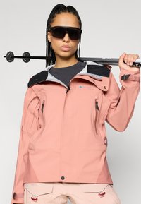 Casaco impermeável rosa claro com detalhes pretos, bolsos com fecho, punhos ajustáveis e detalhes refletivos. O modelo segura um bastão de trekking.