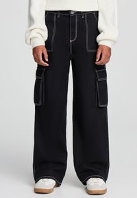 Czarne spodnie cargo o szerokich nogawkach, z białymi przeszyciami, dwiema bocznymi kieszeniami i luźnym fasonem. Noszone z białymi sneakersami.