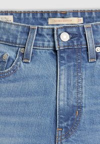 Gros plan sur la taille d'un jean en denim bleu avec bouton en métal, passants de ceinture et étiquette indiquant "725 Kick Bootcut 27."