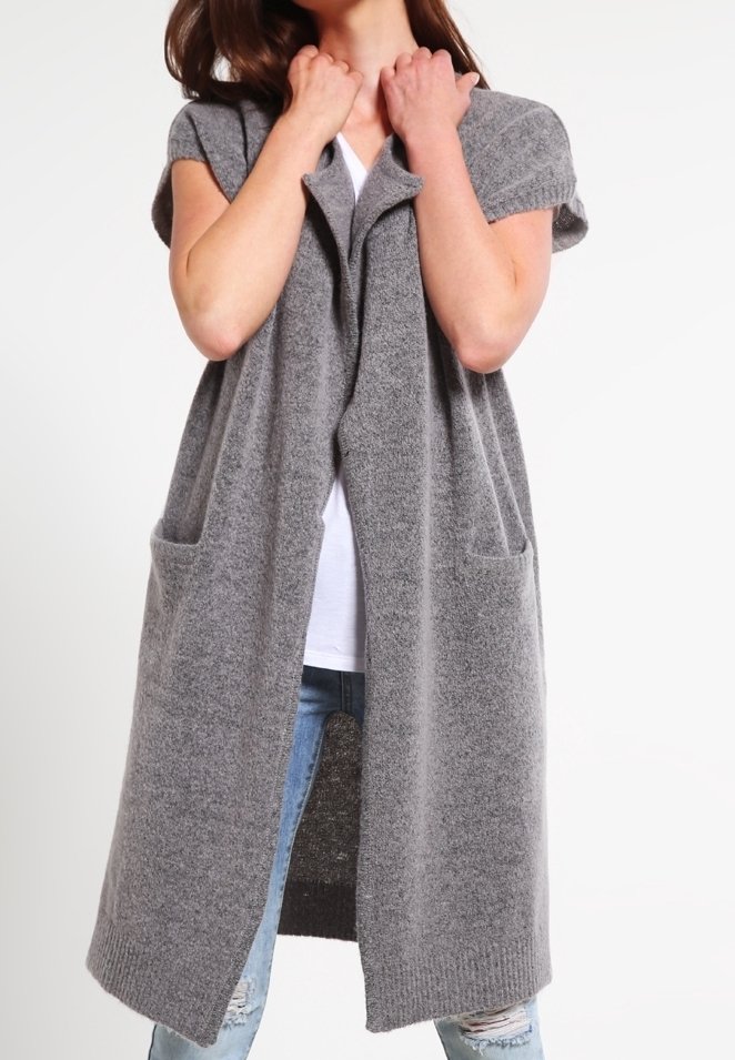 Femme portant un long cardigan gris tricoté sans manches avec des poches, par-dessus une chemise blanche et un jean bleu déchiré.