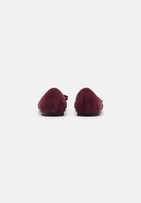 Mocassins en daim bourgogne avec une semelle plate, présentant un bout arrondi et un détail de nœud décoratif sur le dessus pour une texture supplémentaire.