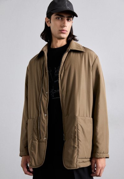 Chaqueta acolchada marrón con diseño de cuello, que cuenta con dos bolsillos frontales y una textura suave. Llevada sobre una camiseta negra, con una gorra negra.