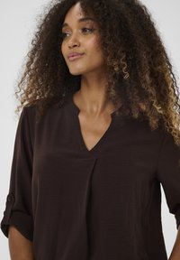 Pruun, kerge pluus V-kaeluse ja lühikeste varrukatega, millel on peen tekstuur ja esiküljel voltdetail.