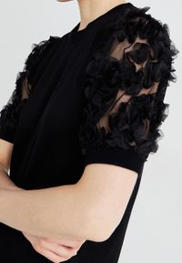 Musta lyhythihainen paita, jossa on teksturoitu läpikuultava kangas ja mustilla kukkaprismaatiikalla koristeltu pinta. Silkkinen puuvillakudos vartalon kohdalla ja resorinen pääntie.