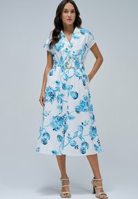 Robe à boutons florale blanche avec des motifs bleus, ceinture avec une boucle en bois ronde, manches courtes évasées et une jupe fluide de longueur mi-mollet.