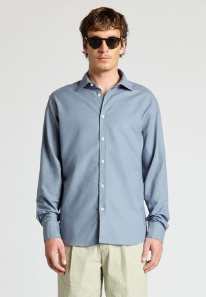 Homme portant des lunettes de soleil noires, une chemise bleue à manches longues boutonnée et un pantalon beige rayé, debout devant un fond clair uni.