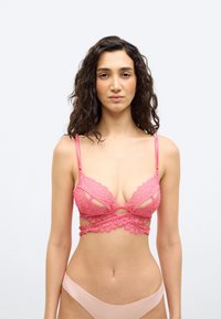 Γυναίκα με σγουρά μαλλιά που φοράει ροζ δαντελωτό σουτιέν με διασταυρούμενους ιμάντες και γυμνό εσώρουχο, στέκεται μπροστά σε απλό λευκό φόντο.