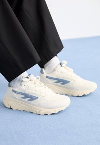 Hvite joggesko med blå detaljer, overdelen i mesh og tekstil, tykk såle og rund form; brukt med lyse sokker og svarte bukser.