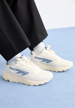 Weiße Sportschuhe mit blauen Akzenten, Obermaterial aus Mesh und Textil, dicke Sohle und abgerundete Form; getragen mit hellen Socken und schwarzen Hosen.