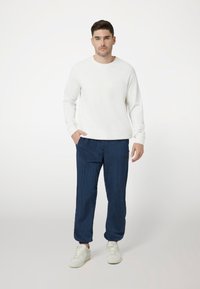 Felpa bianca con testo in rilievo, abbinata a pantaloni blu scuro testurizzati e sneakers bianche. Il modello sta in piedi con una mano in tasca.