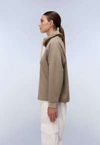 Felpa con cappuccio marrone e finitura testurizzata, con coulisse gialla. Indossata con pantaloni cargo bianchi, modello posato di profilo.
