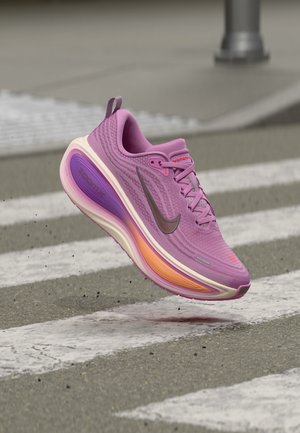 Fialová bežecká obuv Nike s oranžovo-fialovou podrážkou vo vzduchu nad priechodom pre chodcov na ulici.