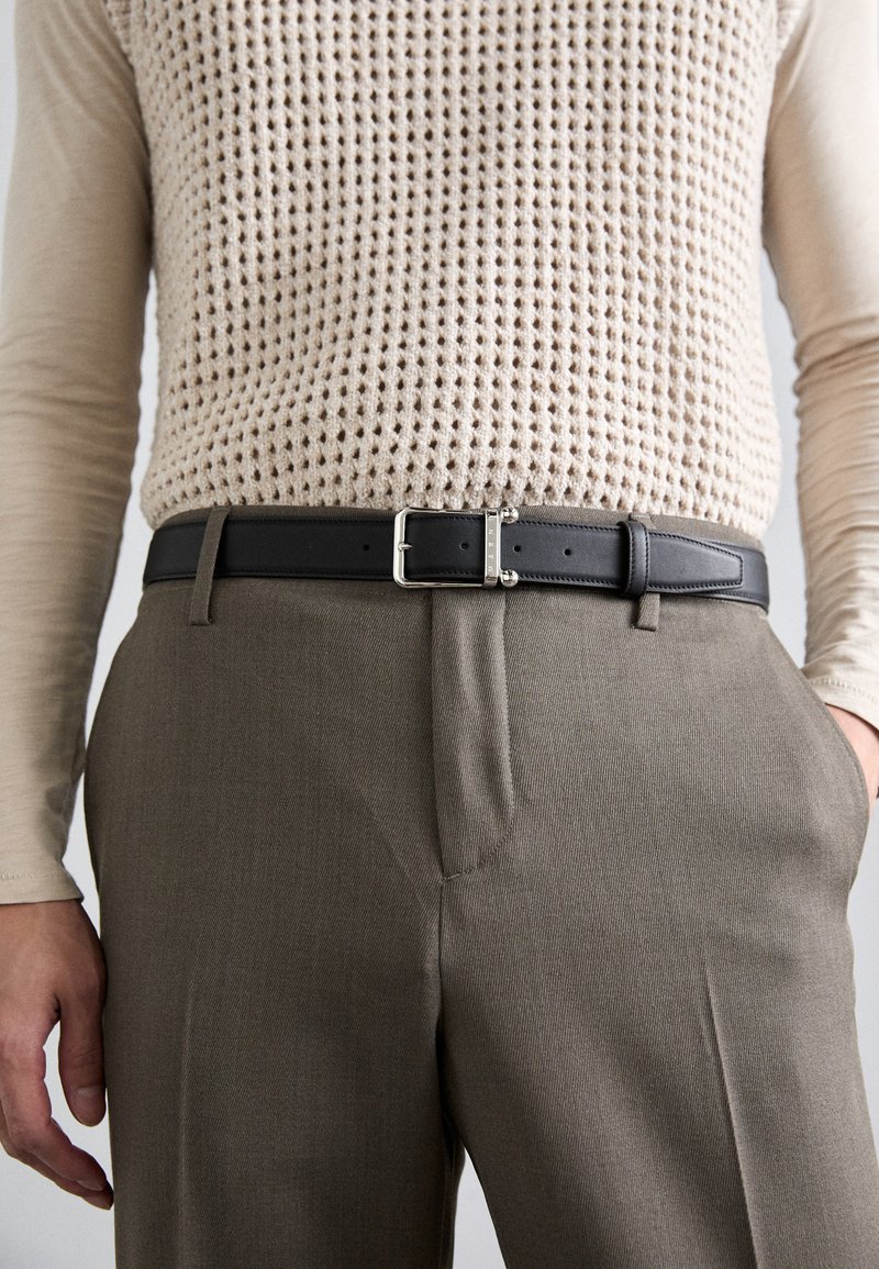 Beige lange mouw top met een wafelgebreide vest, in hoge taille bruine broek gestopt met een zwarte leren riem, een hand in de zak.