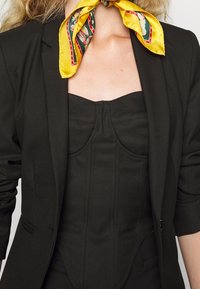 Veste noire avec un corset noir ajusté en dessous. Un foulard en soie jaune à motifs floraux et rayés noué autour du cou.