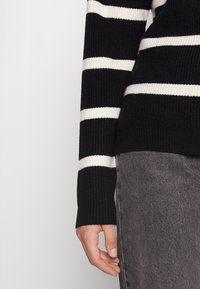 Bras droit et main d'une personne portant un pull noir à rayures blanches et un pantalon en denim gris sur un fond clair uni.