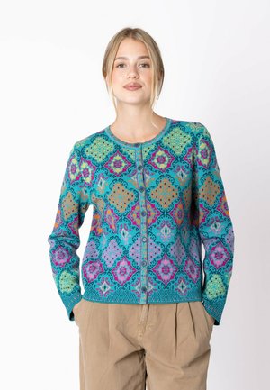 Giovane donna che indossa un cardigan colorato a motivi con bottoni e pantaloni beige, in piedi con le mani nelle tasche su sfondo bianco semplice.