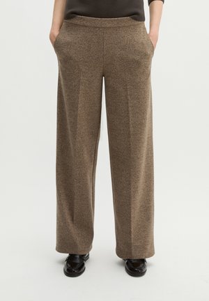 Pantalon droit en laine marron porté avec des chaussures noires, mains dans les poches, sur un fond clair uni.