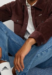 Brun kordjacka med knappstängningar, tillsammans med ljusblå denimjeans och vita strumpor med en svart logotyp.