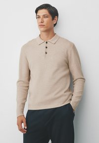 Mand iført en beige langærmet skjorte med krave og tre knapper, mørke bukser, stående mod en hvid væg, en hånd i lommen.