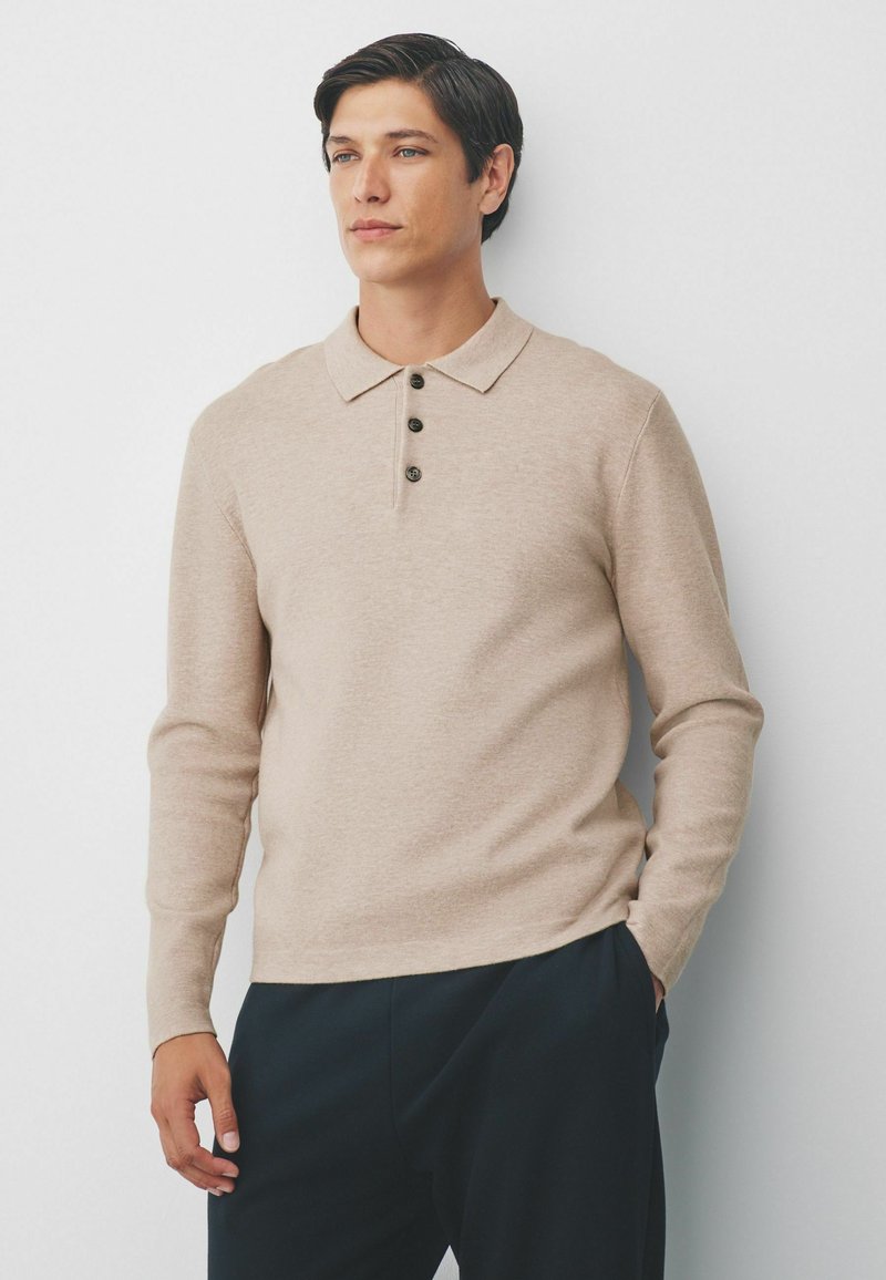 Mand iført en beige langærmet skjorte med krave og tre knapper, mørke bukser, stående mod en hvid væg, en hånd i lommen.