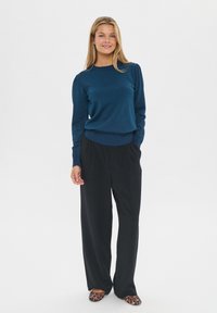 Fiatal nő áll, kék hosszúujjú pulóvert, laza fekete nadrágot és leopárdmintás lapos cipőt visel, egy sima fehér háttér előtt.