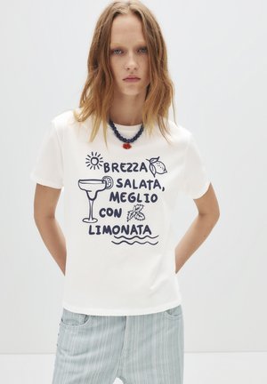 T-shirt bianco in cotone con testo e grafiche stampate in blu, inclusi un bicchiere di cocktail, un limone e illustrazioni di sole. Pantaloni a righe azzurri.