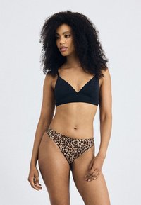 Femeie cu păr creț, îmbrăcată cu un bralet negru și bikini cu imprimeu de leopard, stând pe un fundal luminos simplu, privind spre dreapta.