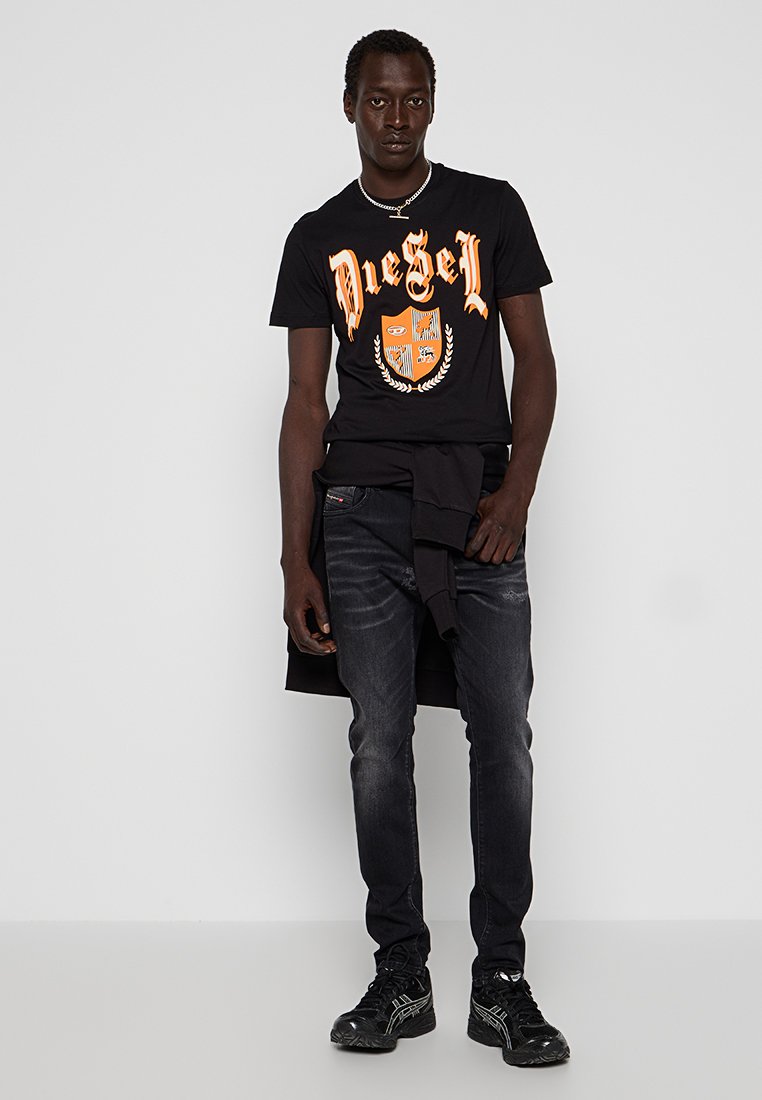 Mann trägt ein schwarzes Diesel-T-Shirt mit orangefarbenem Logo, schwarze Jeans, schwarze Sneakers und einen schwarzen Pullover um die Taille gebunden, steht vor einer weißen Wand.