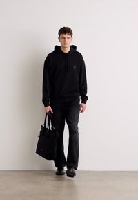 Schwarzer Hoodie mit einem kleinen blauen Logo, kombiniert mit dunklen Jeans und einer schwarzen Tragetasche. Der Stoff wirkt glatt und entspannt.