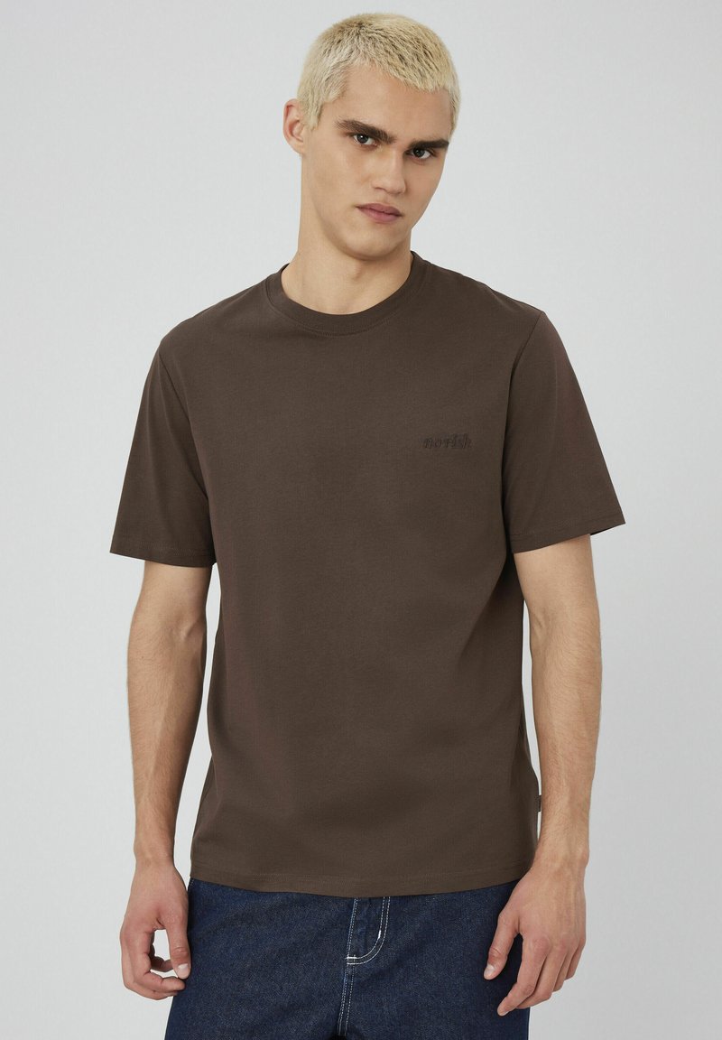 T-shirt en coton marron avec col rond, manches courtes et petit logo brodé sur la poitrine, associé à un jean en denim bleu.