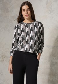 Langemouwen top in een geometrisch zwart, grijs en wit patroon, gemaakt van zachte stof, gecombineerd met zwarte broeken met zakken.